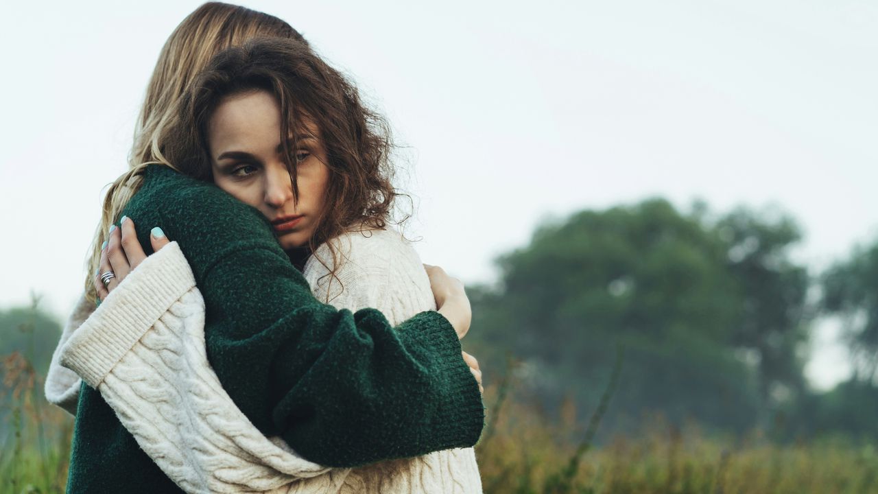 Жінка з сумним та підозрілим поглядом обіймає подругу, ілюструючи приховані ознаки заздрості та нещирої підтримки.