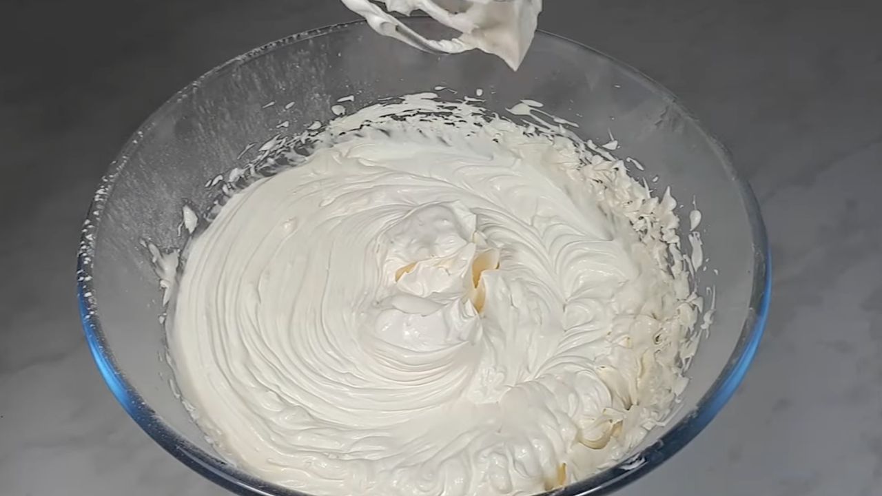 Густий та шовковистий сметанний крем у процесі збивання міксером у скляній мисці.