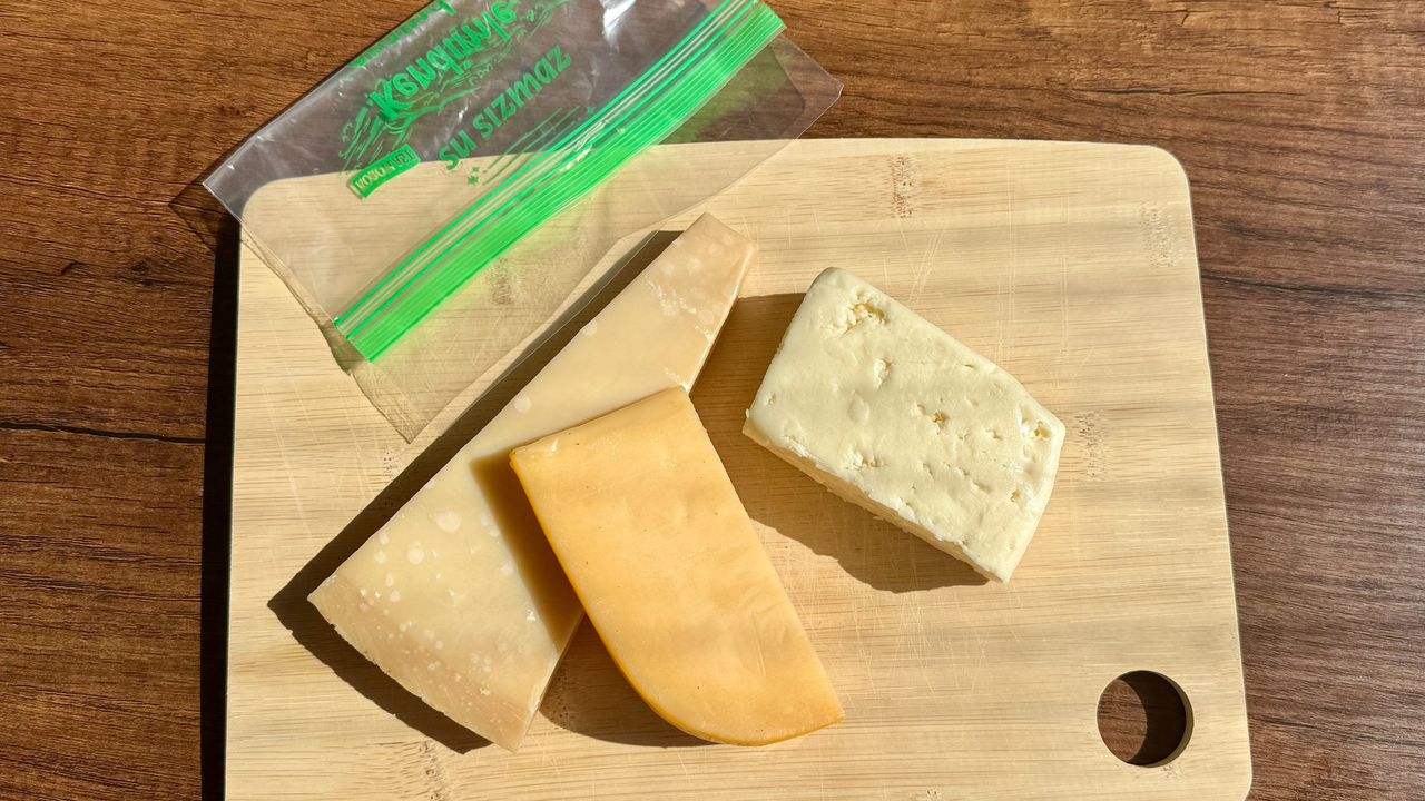 Три види твердого сиру на дерев'яній дошці та зип-пакет для тривалого зберігання продуктів свіжими.